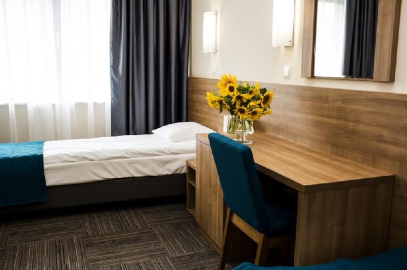 Łóżko pod oknem z szafką nocną, biurkiem i wazonem ze słonecznikami.