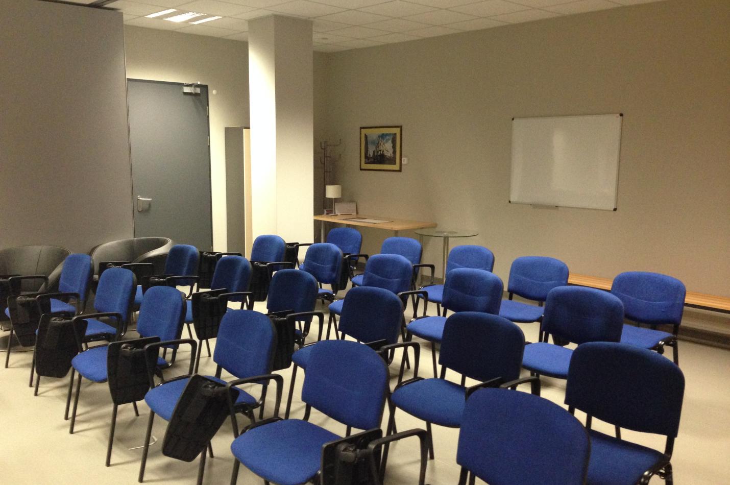 Sala szkoleniowa z rzędem niebieskich krzeseł i tablicą na ścianie.