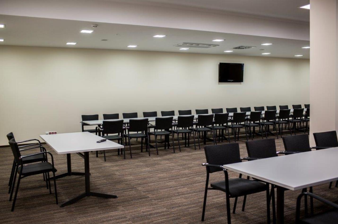 Sala konferencyjna z długimi stołami, rzędem krzeseł i telewizorem na ścianie.