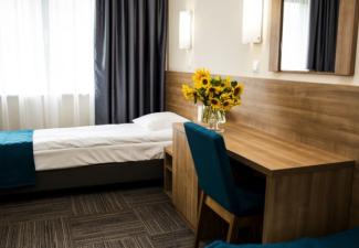 Łóżko pod oknem z szafką nocną, biurkiem i wazonem ze słonecznikami.