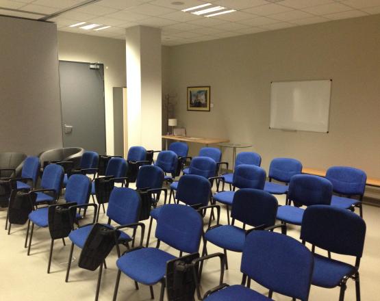 Sala szkoleniowa z rzędem niebieskich krzeseł i tablicą na ścianie.