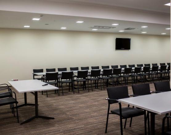 Sala konferencyjna z długimi stołami, rzędem krzeseł i telewizorem na ścianie.
